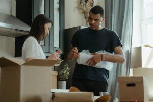 Man and a woman standing next to a pile of cardboard boxes and trying to pack fragile items for their move