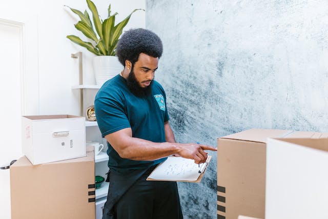 Moving Pros will carefully and efficiently pack and unpack your belongings. Alt text: Man with a notepad, standing between cardboard boxes