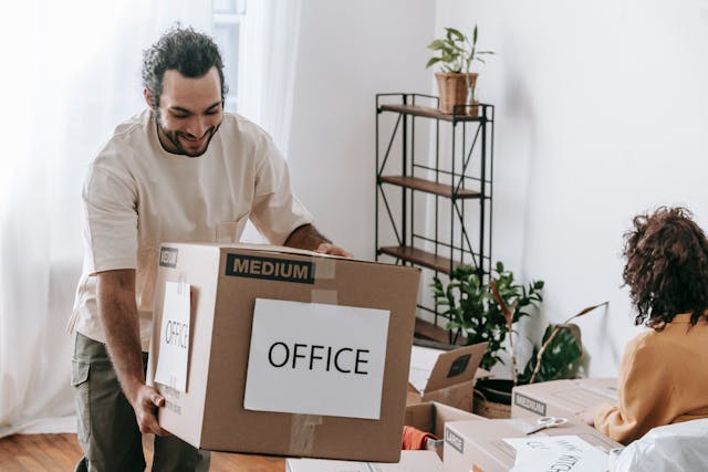 Two people packing office supplies into a labeled box marked “OFFICE” in a room filled with moving boxes